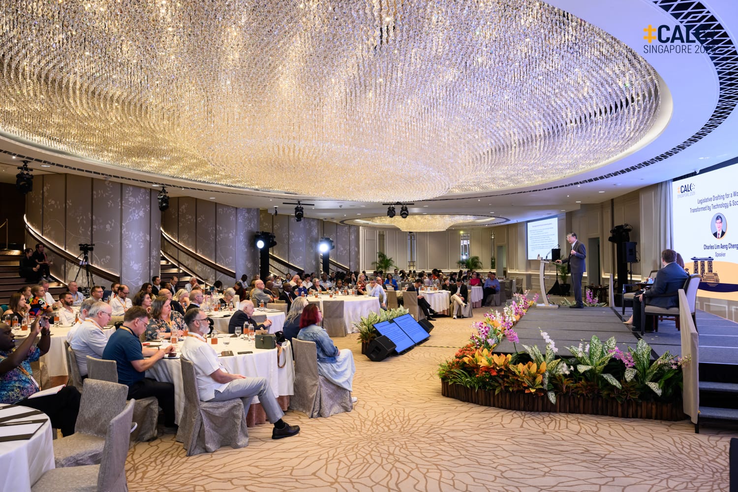 A wide shot of the ballroom at The Fullerton Hotel Singapore during a Conference session, with speaker Charles Lim Aeng Cheng presenting at the podium, while Benjamyn McKay and Kerri Sinclair look on seated on stage.